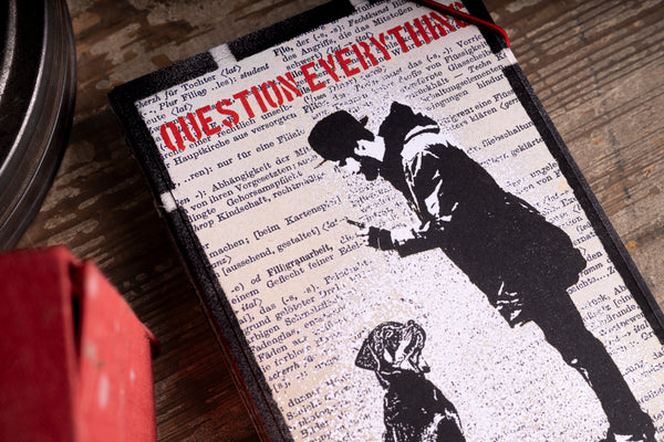 A close-up of notebook, featuring a black-and-white graphic of a person and dog over a dictionary-print background, with bold red text reading 'QUESTION EVERYTHING' and a thin red string tied at the top, placed on a wooden surface.