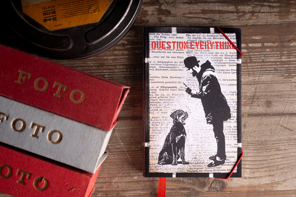 A notebook titled 'QUESTION EVERYTHING' with a black-and-white graphic of a man and dog on a newspaper-style background, styled on a wooden surface next to film reels and books.
