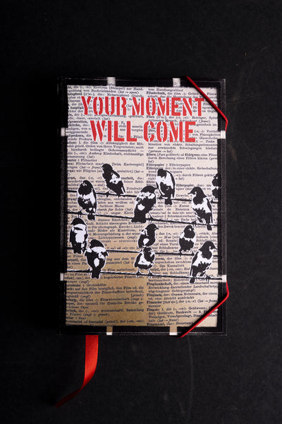 A rectangular handmade notebook featuring a vintage dictionary page background with the red text 'YOUR MOMENT WILL COME' and black bird silhouettes, displayed on a dark background with a red ribbon bookmark.