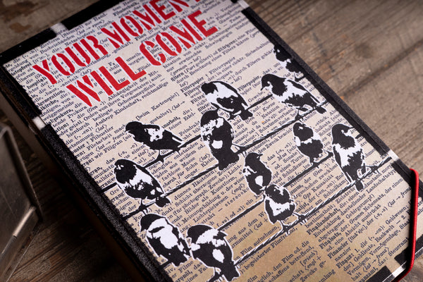 A hardcover handmade notebook lying on a rustic wooden surface. The cover features a vintage design with a black and white bird stencil pattern over a beige book page background, with the bold red text 'YOUR MOMENT WILL COME' at the top.