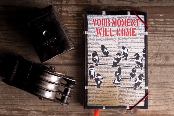 A rectangular  handmade notebook with a vintage-style cover featuring the phrase 'YOUR MOMENT WILL COME' in red text over a background of black-and-white bird illustrations on a book page. The notebook is displayed on a rustic wooden surface alongside an antique pocket watch and a small dark metal box.