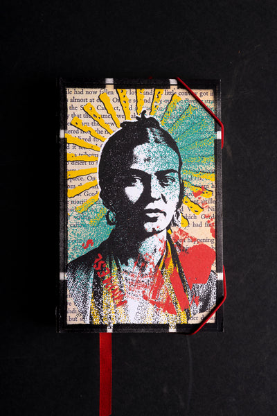 A  journal cover featuring a stylized portrait of a woman in black and white, overlaid with bright yellow, teal, red, and blue graphic elements and text fragments. The item is photographed against a dark black background with a red ribbon bookmark hanging from the bottom.