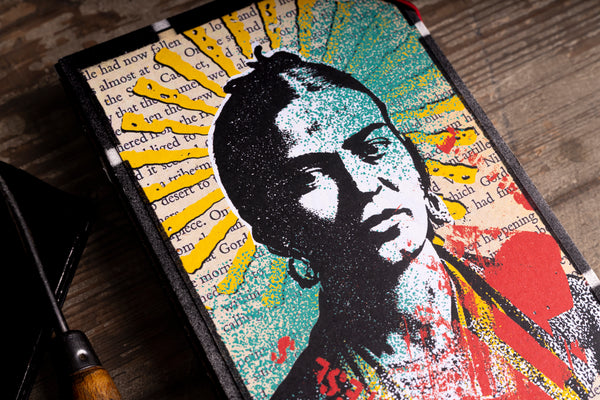 A close-up of a hardcover journal featuring a vibrant pop-art style portrait of Frida Kahlo, printed over a background of vintage book pages. The cover has a black textured border and is shown resting on a rustic wooden surface.