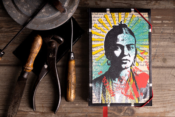 A notebook with  stylized portrait of a woman with a yellow sunburst background, styled on a rustic wooden table alongside vintage tools.