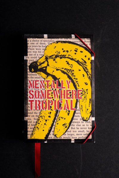 A rectangular handmade journal  with a red ribbon bookmark, featuring a graphic cover design of yellow bananas over a vintage book page background with the text 'MENTALLY SOMETIME TROPICAL' in red and black.