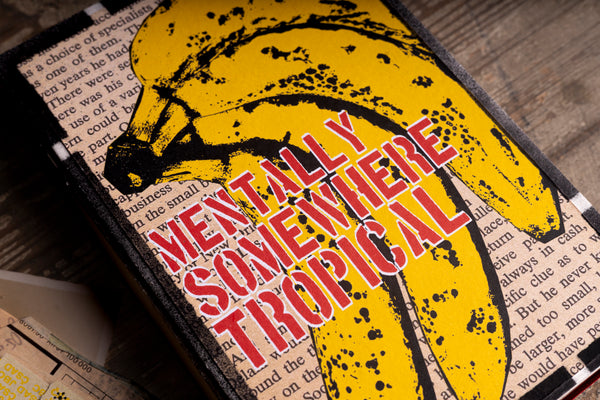 A close-up of a notebook cover featuring a yellow banana illustration with black splatter details and the title 'MENTALLY SOMEWHERE TROPICAL' in red and white text, placed on a wooden surface.