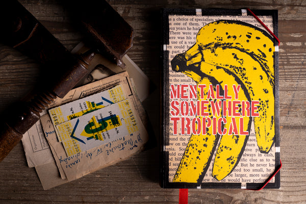 A hardcover handmade notebook titled 'MENTALLY SOMEWHERE TROPICAL' featuring a yellow banana graphic over a newspaper print background, styled on a rustic wooden table next to a wooden spoon and vintage paper.