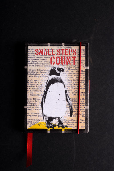 Handmade notebook "Penguin" with a screen-printed cover of a black and white penguin standing on a yellow mound over German dictionary text, under the red stencil words "SMALL STEPS COUNT". It has a black textured border, a red elastic closure, a red ribbon bookmark and a pen loop, on a black background.