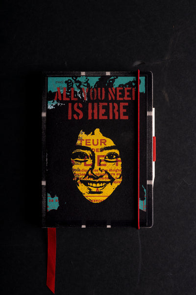 Handmade notebook "Power Girl" with a screen-printed cover in black, yellow, teal and red, showing a smiling yellow face over French and German printed text under the stencil words "ALL YOU NEED IS HERE". It has a red elastic closure, a red ribbon bookmark and a pen loop, on a black background.

