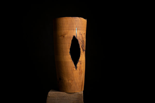 A natural wood cylindrical object handturned from red oak with a prominent dark heart-shaped  hollow opening, standing upright on a small wooden block against a solid black background.