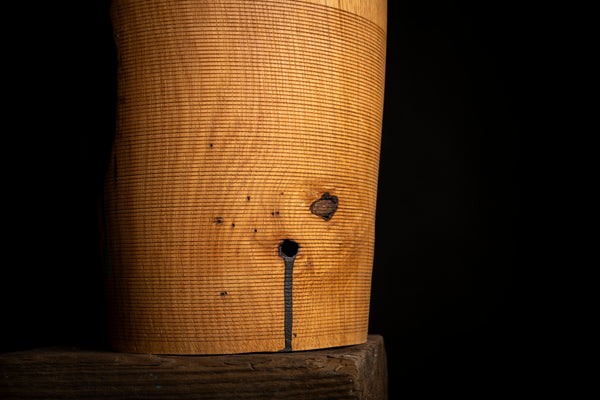 A close-up of a cylindrical wooden object with a prominent natural grain pattern, a small knot, and a dark vertical line near the bottom, set against a solid black background.