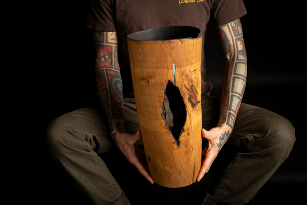 A person with tattooed arms holds a tall, cylindrical wooden object with a large natural fissure and dark interior against a black background. Object is turned on the lathe from red oak. 