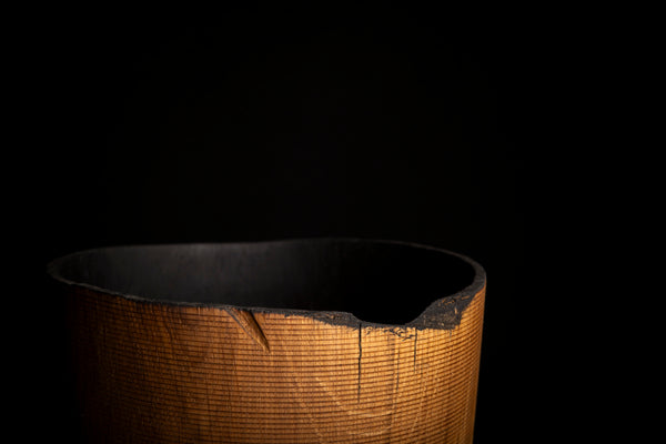 A close-up of a textured, ribbed cylindrical  vase with a warm golden-brown exterior and a dark black interior, set against a solid black background.