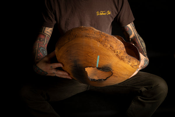 A person seated against a black background holds a large, irregularly shaped natural wood bowl with a prominent live edge and a small blue pencil sticking out of a cutout. Artist work under the name Maalstrom.studio in Prague