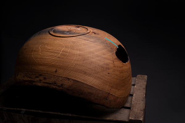 A rustic hand turned oak wooden bowl with a hollow center and a circular cutout on top, displayed on a wooden stand against a dark background