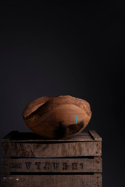 A rustic hand turned wooden bowl with a natural rough edge, displayed on top of stacked wooden crates against a dark studio background.
