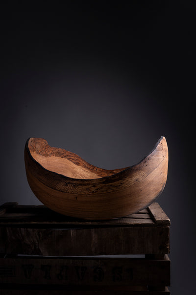 A wooden bowl tuned on the lathe with a curved, crescent-like shape, displayed on a rustic wooden crate against a dark, moody background.