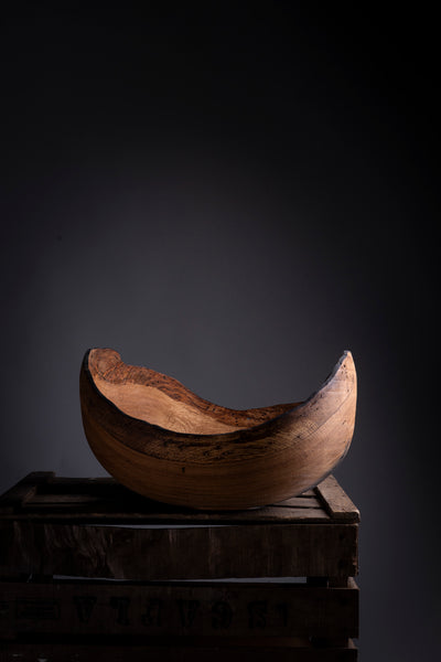 A rustic turned wooden bowl from oak with a curved, crescent-like shape, displayed on a dark wooden crate against a dark gray background.