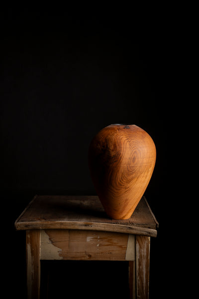 A teardrop-shaped wooden vase with rich grain patterns, displayed on a rustic wooden table against a solid black background.