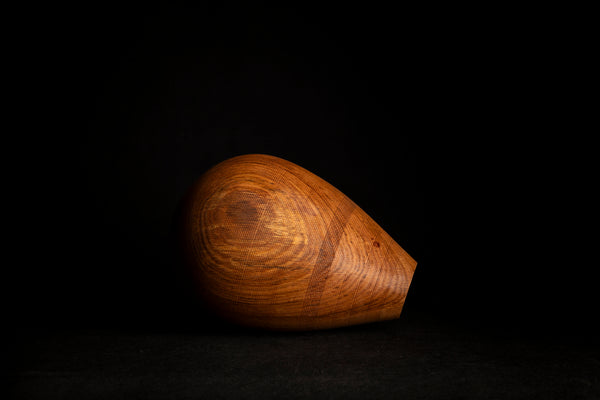 A teardrop-shaped wooden object with a rich warm brown wood grain, photographed on a solid black background. Hand turned in Prague on the lathe from red oak. 