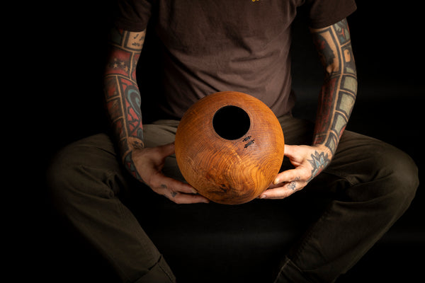 A person with tattooed arms holds a round, wooden vessel with a central hole, set against a solid black background. The vessel is made on the lathe from red oak.