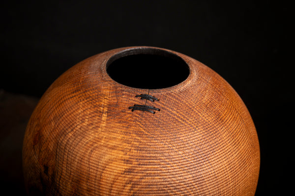 A close-up of a handcrafted wooden vessel with a circular opening at the top, showcasing detailed wood grain and dark stitched accents against a solid black background. Turned from red oak on the lathe by maalstrom.studio