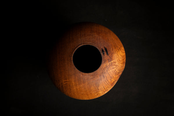 A handcrafted wooden hollow form  with a circular hole in the center, showcasing a rich warm brown wood grain texture, photographed against a dark black background. Turned by maalstrom.studio