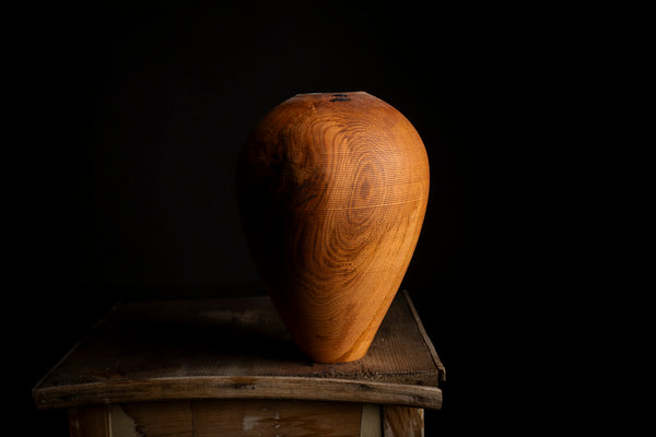 A teardrop-shaped wooden vase with a rich warm brown finish by tung oil and prominent natural wood grain, displayed on a rustic wooden pedestal against a solid black background.