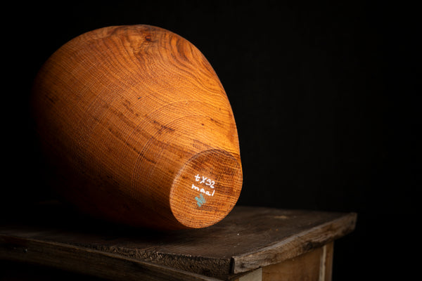 A wooden hollow form with a rounded teardrop shape, resting on a rustic wooden table against a solid black background. The wood has a warm honey-brown finish with visible grain patterns and a small white hand-written marking on the circular end. Turned on the lathe by artist from Prague creating under a name maalstrom.studio