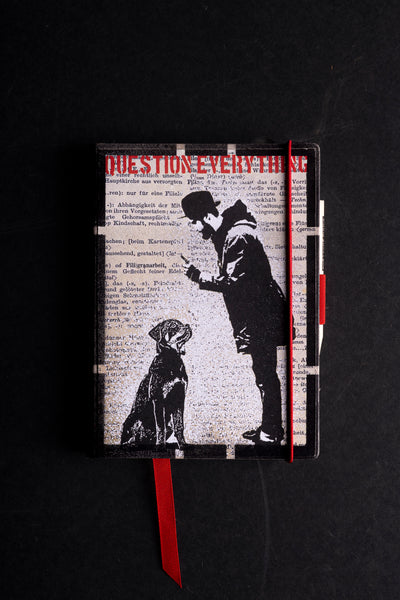 Handmade notebook "The Dog and (Not) His Master" with a black and white screen-printed cover of a man in a hat looking at his phone while a seated dog gazes up at him, over printed German dictionary text, under the red stencil words "QUESTION EVERYTHING". It has a black textured border, a red elastic closure, a red ribbon bookmark and a pen loop, on a black background.