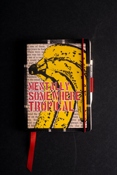 Handmade notebook "Tropical Banana Soul" with a screen-printed cover of a large yellow banana bunch over printed book-page text, with the red stencil words "MENTALLY SOMEWHERE TROPICAL". It has a black textured border, a red elastic closure, a red ribbon bookmark and a pen loop, on a black background.
