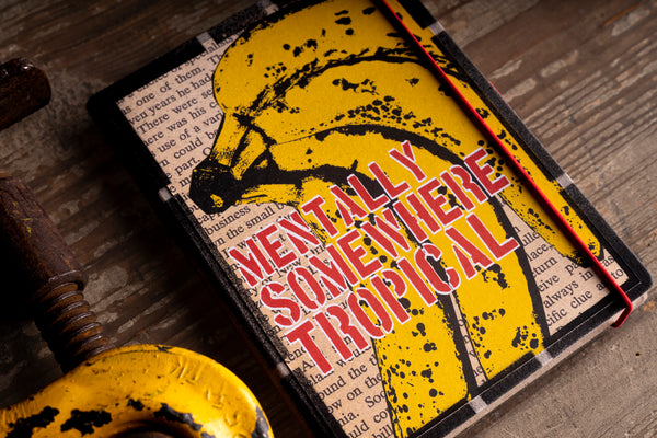 Angled close-up of the "Tropical Banana Soul" handmade notebook cover, showing the yellow screen-printed bananas with black speckles, the red stencil lettering "MENTALLY SOMEWHERE TROPICAL" outlined in white, and the red elastic band, beside a rusty vintage clamp on weathered wood.
