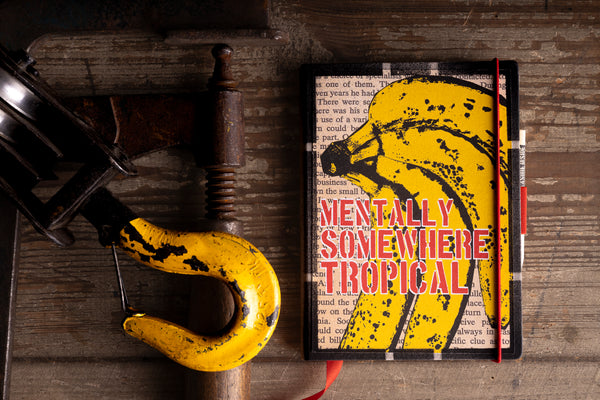 Handmade notebook "Tropical Banana Soul" with a bold yellow banana cover and red stencil text "MENTALLY SOMEWHERE TROPICAL", placed beside a yellow-painted industrial hook and rusty clamp on a rustic wooden surface.