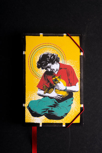 A hardcover handmade journal with a black cover, featuring a vibrant pop-art style illustration of a woman in a red shirt and teal pants holding a yellow cat, set against a bright yellow background. The notebook is photographed flat on a black surface, secured with white tape at the corners and a red ribbon bookmark hanging from the bottom.