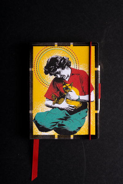 Handmade notebook "Woman with Cat on Lap" with a screen-printed cover of a woman in a red shirt and teal trousers holding a Siamese cat, framed by a dashed red, black and teal halo on a yellow background. It has a black textured border, a red elastic closure, a red ribbon bookmark and a pen loop, on a black background.