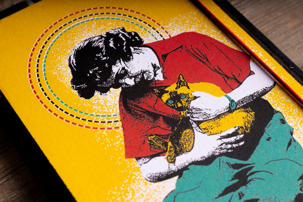 A close-up of a colorful handmade notebook design showing a stylized illustration of a person in a red shirt hugging a yellow cat, with concentric dotted circles behind them, photographed on a wooden surface.