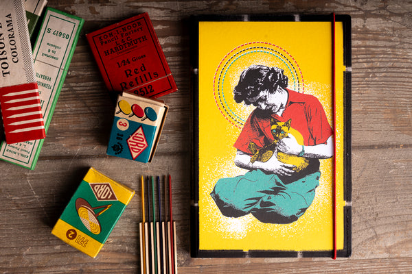A black hardcover notebook with a vibrant yellow cover featuring a graphic illustration of a woman holding a cat, styled on a rustic wooden table alongside vintage matchboxes and colored pencils.