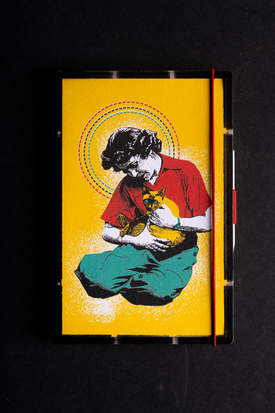 A hardcover notebook with a bright yellow cover featuring a stylized pop-art illustration of a woman in a red shirt and teal pants on a black background holding a cat on her lap