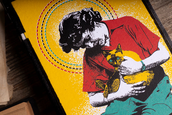 A close-up of a colorful pop-art style print on notebook cover showing a woman in a red shirt holding a yellow cat, with concentric dotted circles and a bright yellow background.