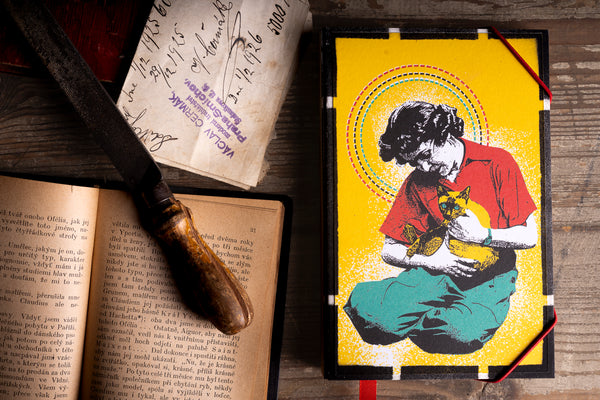 A handmade notebook featuring a stylized illustration of a woman holding a cat, displayed on a rustic wooden table alongside an open book, a vintage knife, and handwritten notes.