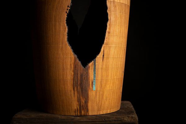 A close-up of a cylindrical wooden object with a large irregular black hole or opening on the front, showing a warm honey-brown wood grain surface of red oak and a small vertical blue accent, set against a black background.