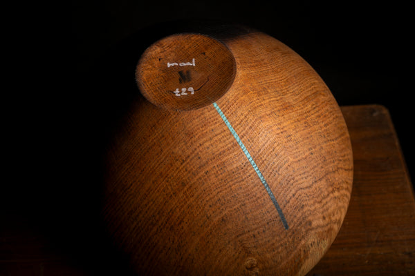 A close-up of a wooden vessel shown upside down on a dark surface, with visible wood grain and a small painted blue-green line running diagonally across the base.